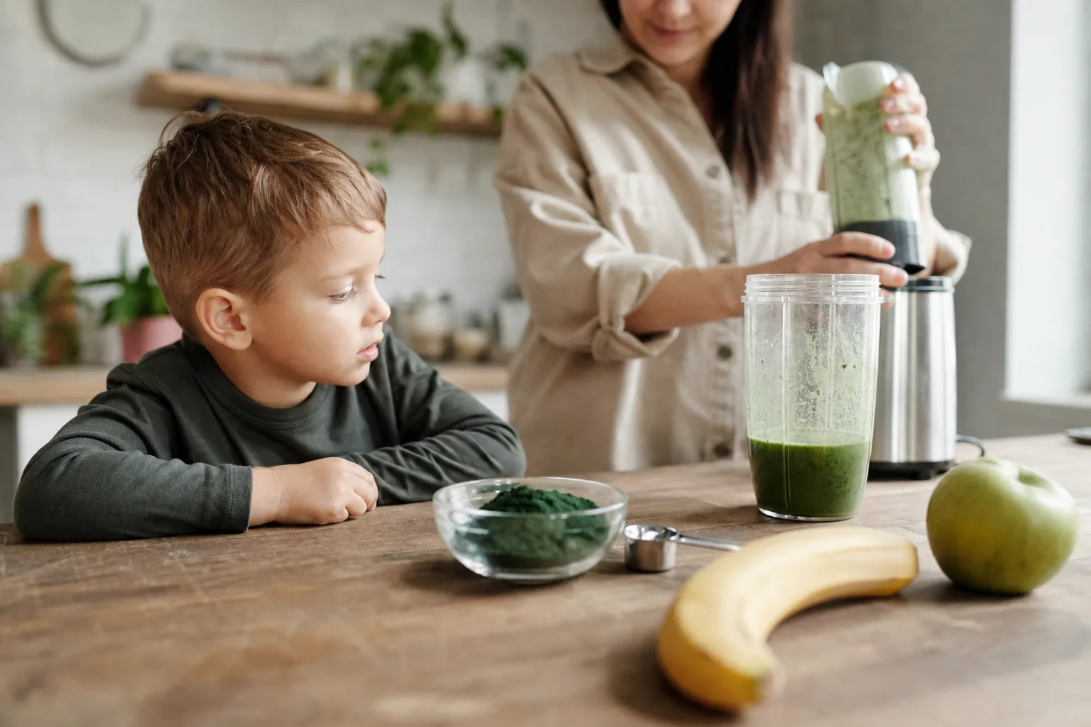 Konyhaasztalon egy kis üvegtálban élénkzöld spirulina por, mellette banán, alma, mérőkanál és egy félig megtöltött turmixpohár; a szülő a pultnál turmixot készít, miközben a gyerek kíváncsian nézi a zöld port.