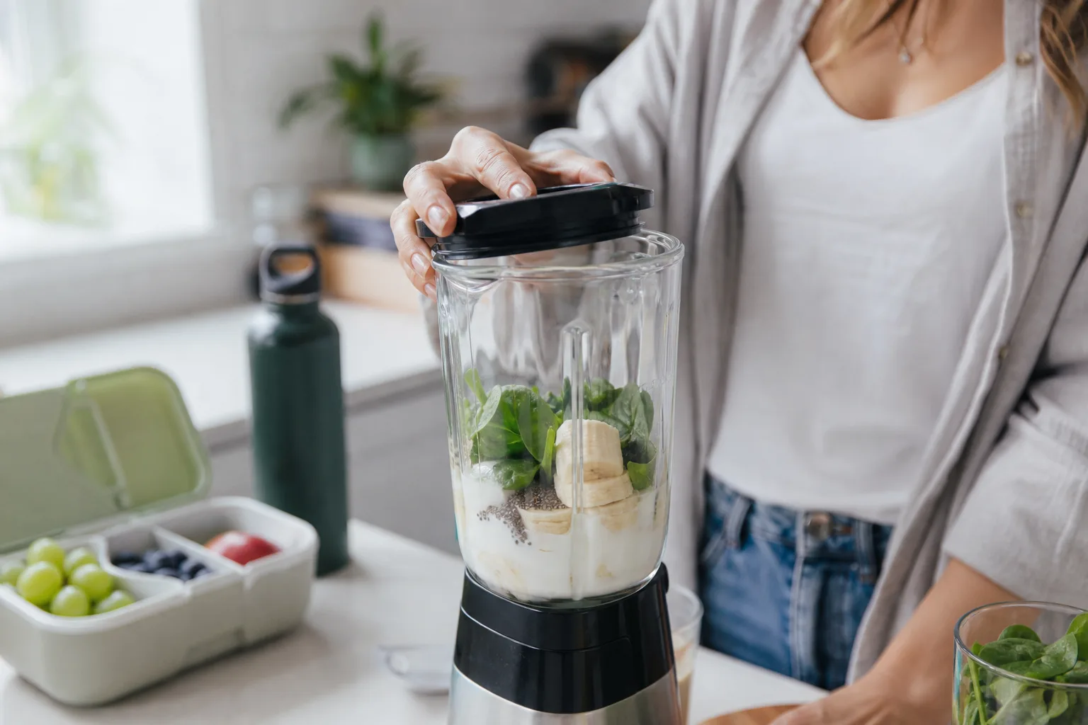 Reggeli konyhában turmixgép poharába banánkarikák, spenótlevelek, joghurt és egy kanál chia mag kerül; a háttérben uzsonnásdoboz gyümölccsel és egy kulacs, miközben valaki az egyik kezével a turmixgép fedelét helyezi fel.