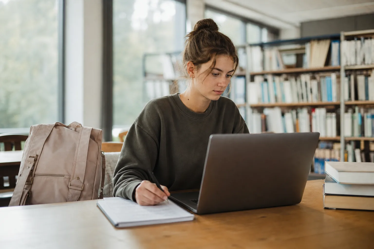 női diák ül könyvtárban, hátizsák az asztalon, jegyzeteket készít laptopon