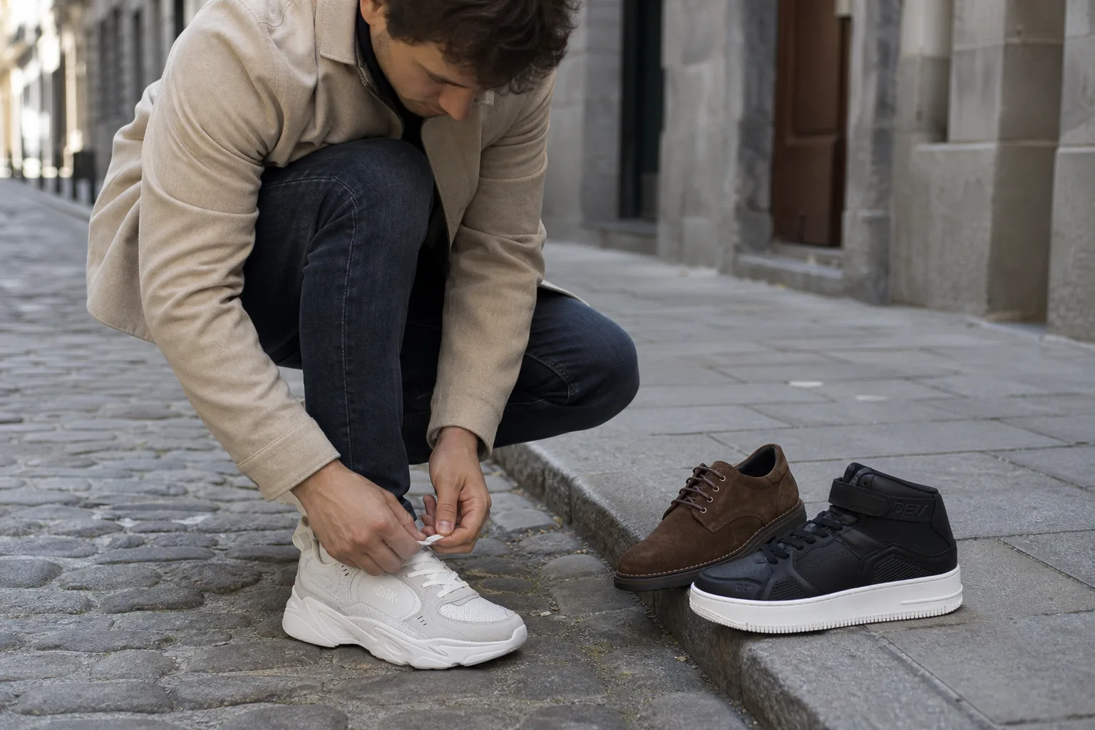 Egy férfi macskaköves utcán áll sötét farmerben és bézs kabátban, az egyik lábán letisztult fehér bőr sneaker, mellette a járdán egymás mellett egy barna velúr derby és egy fekete vászon tornacipő, miközben lehajolva a sneaker fűzőjét köti.