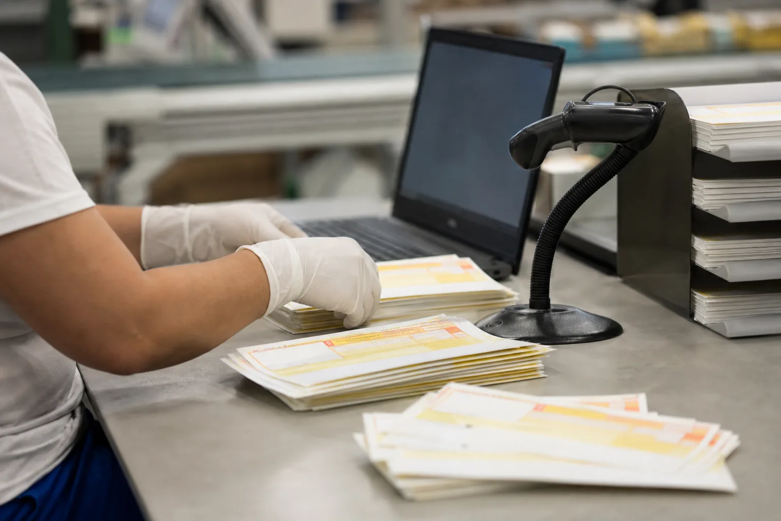 Egy nyomdai előkészítő asztalon üres sárga-fehér csekknyomtatványok, mellette laptop képernyője elfordítva, vonalkódolvasó és iratrendező tálca; egy operátor gumikesztyűben külön kupacba rendezi a megszemélyesítésre váró csekkeket.
