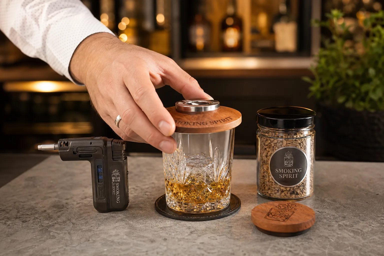 Konyhapulton elhelyezett whiskeyspohár nagy jégkockával, mellette fa koktélfüstölő fedél, kis konyhai flambírozó és zárt üveg tégelyben faforgács; egy kéz éppen a füstölő fedelet igazítja a pohár tetejére, a háttérben tiszta, rendezett otthoni bárpult látszik.