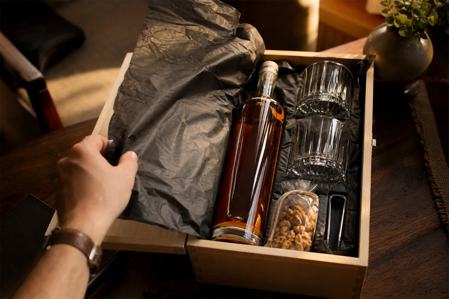 Fa étkezőasztalon nyitott, natúr fadobozban egy címke nélküli whiskey-palack, két vastag talpú pohár, jégcsipesz és egy kis zacskó pörkölt dió; egy kéz éppen a selyempapírt hajtja félre a palack elől, meleg délutáni fényben.