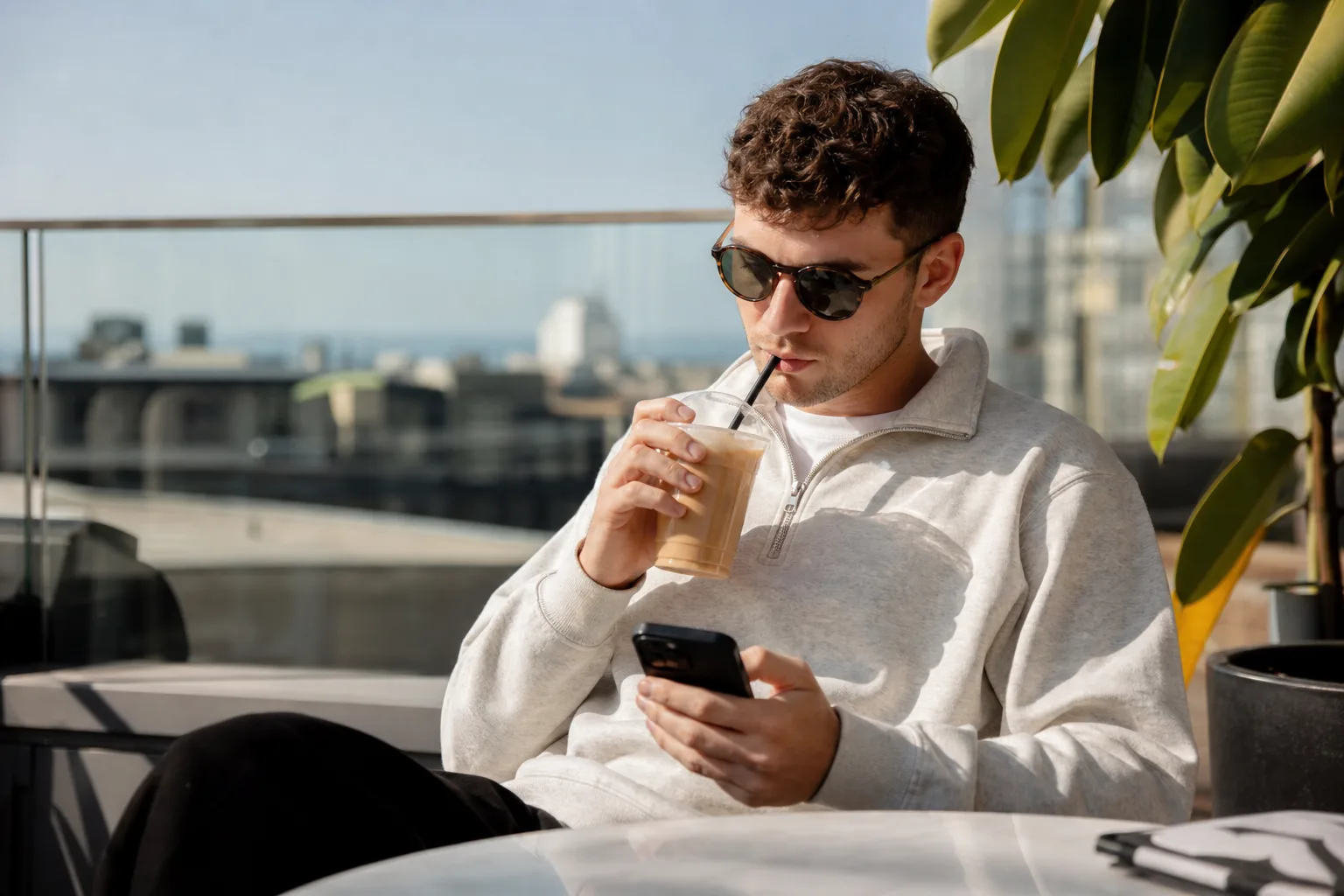 Egy ember ül egy teraszon, napszemüveget visel, és egy melegítő felsőt hord, miközben egy jégkávét iszik és a telefonján böngészik.