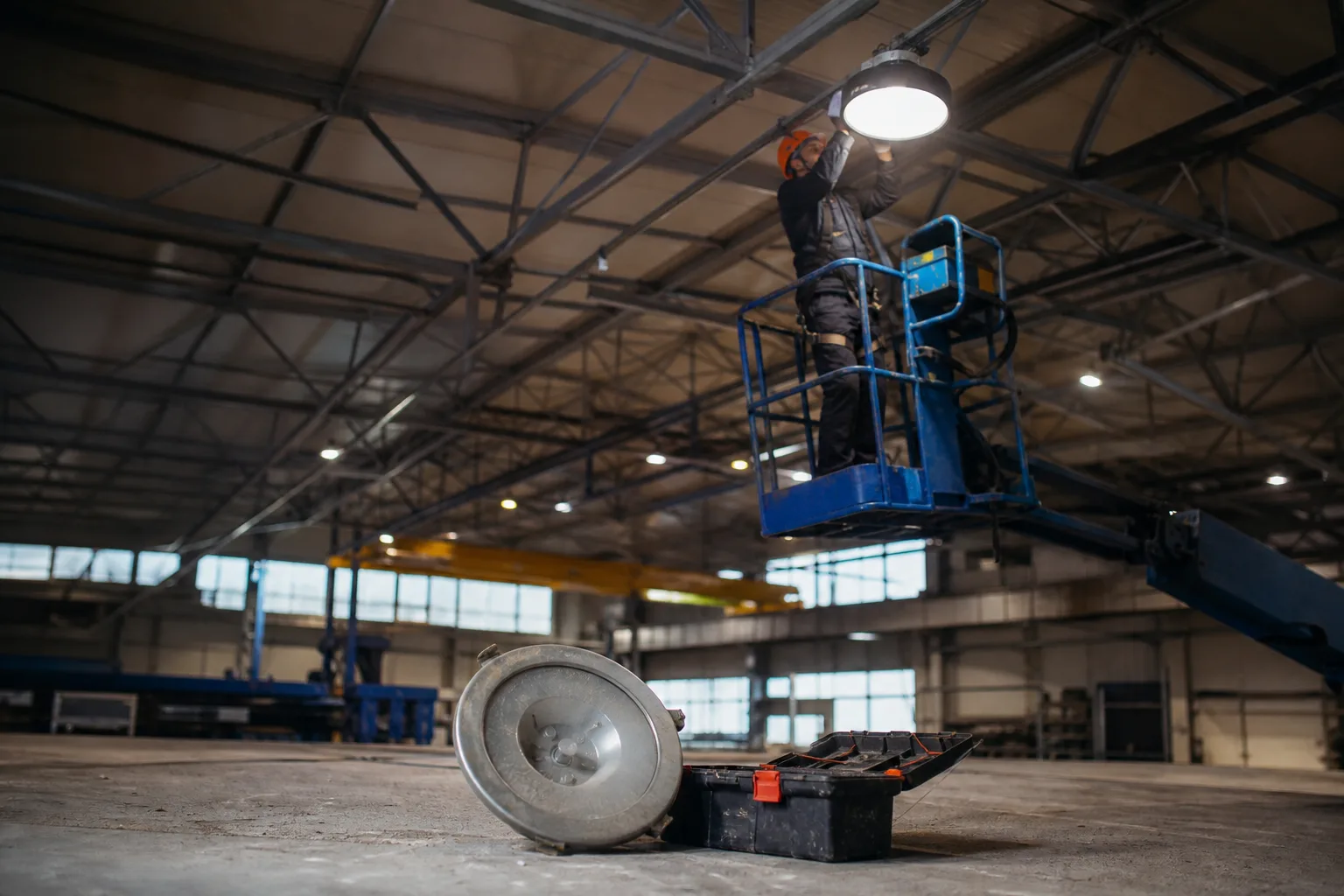 Egy ipari csarnok acélgerendái alatt egy szerelő kosaras emelőből kerek LED high-bay csarnokvilágítót rögzít a tartósínre, miközben a padlón egy régi, poros nagyméretű lámpatest fekszik a szerszámosláda mellett.