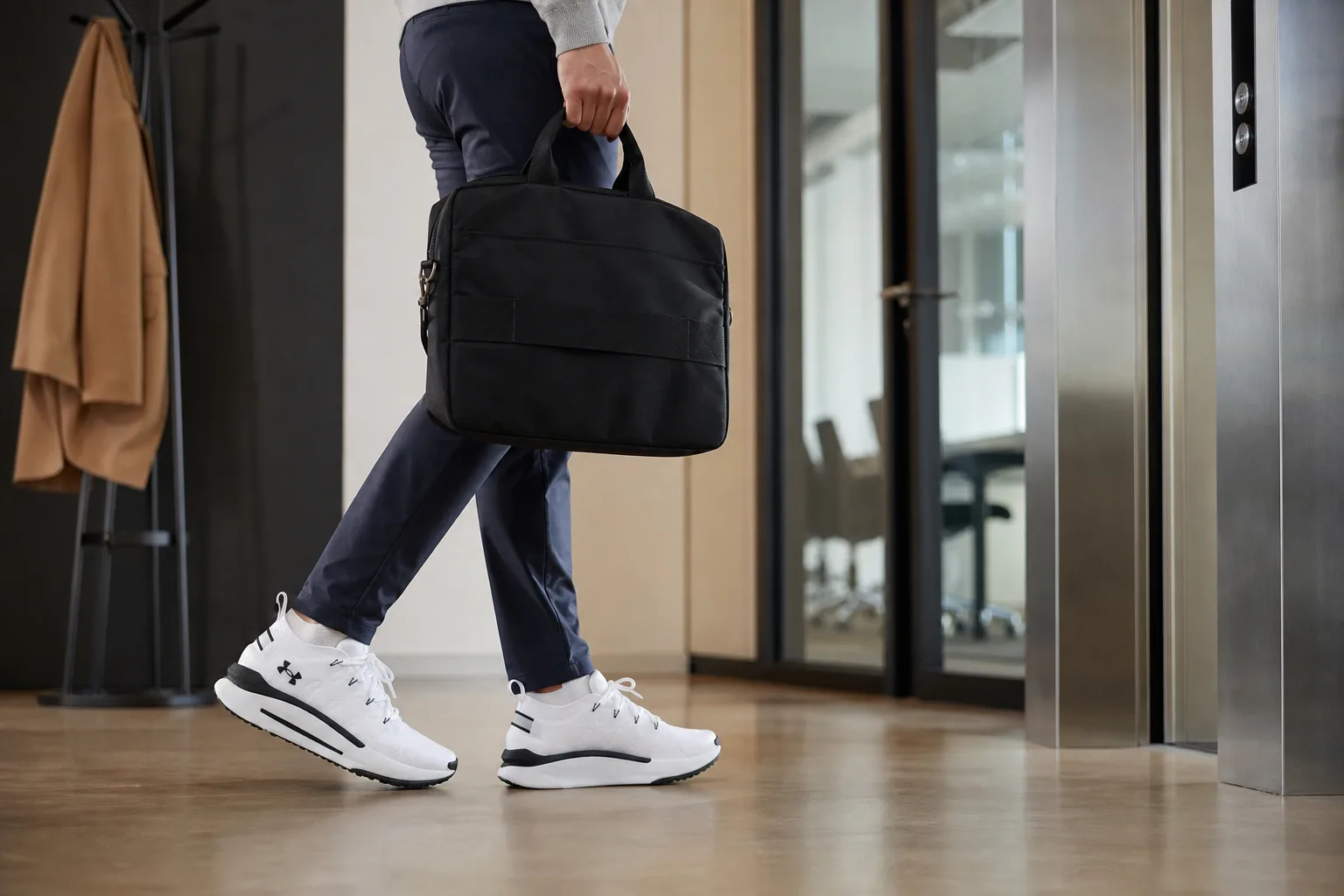 Irodai folyosón egy ember visszafogott sportcipőben lép a lift felé, egyik kezében laptop táska, a háttérben kabátakasztó és zárt tárgyalóajtó; a cipő jól látható a sima padlón járás közben.