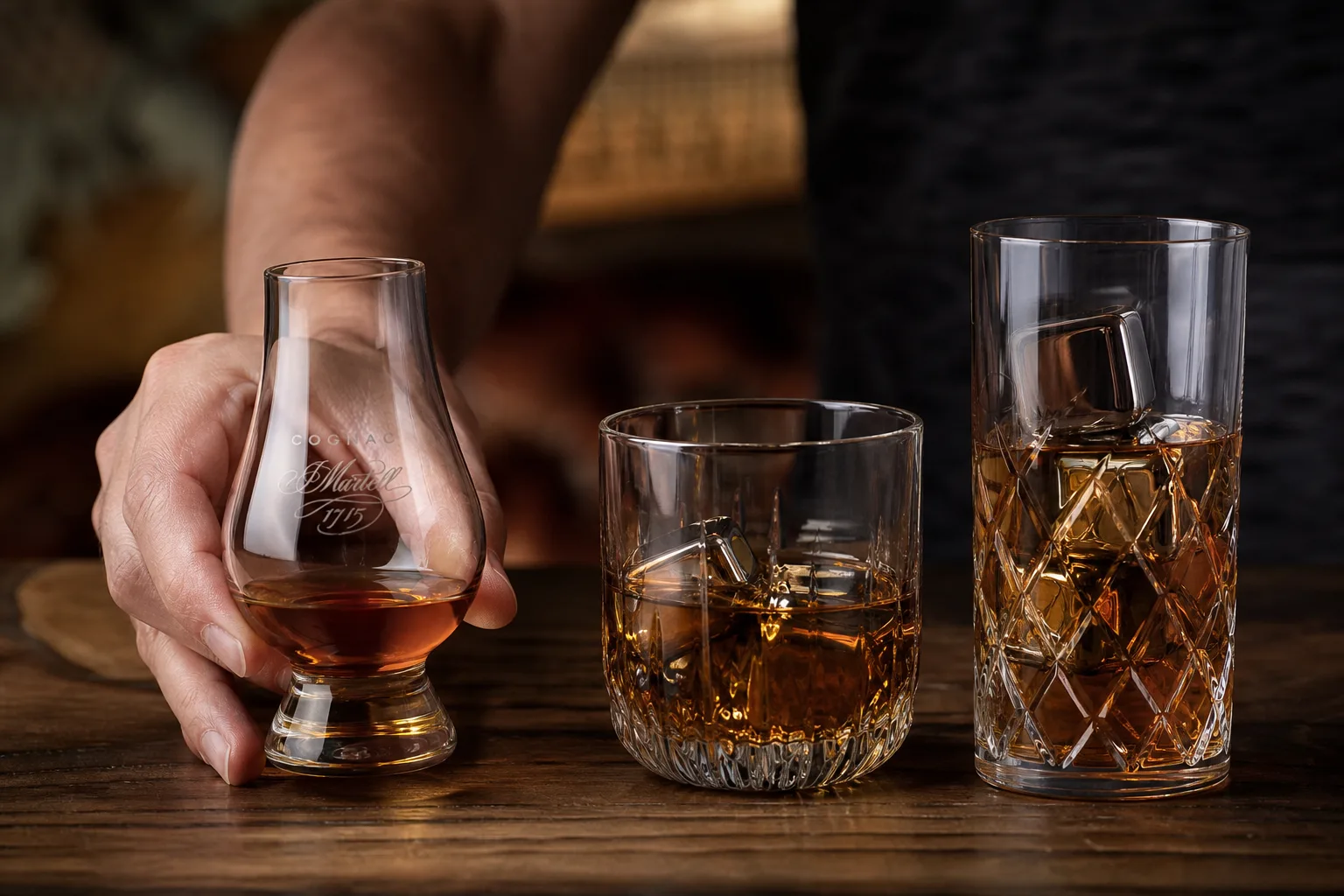 Sötét diófa asztalon három különböző whiskey pohár áll egymás mellett: tulipán formájú kóstolópohár kevés borostyánszínű whiskeyvel, széles old fashioned pohár nagy jégkockával, és magas highball pohár jéggel; egy kéz éppen a tulipánpoharat emeli ki a sorból.