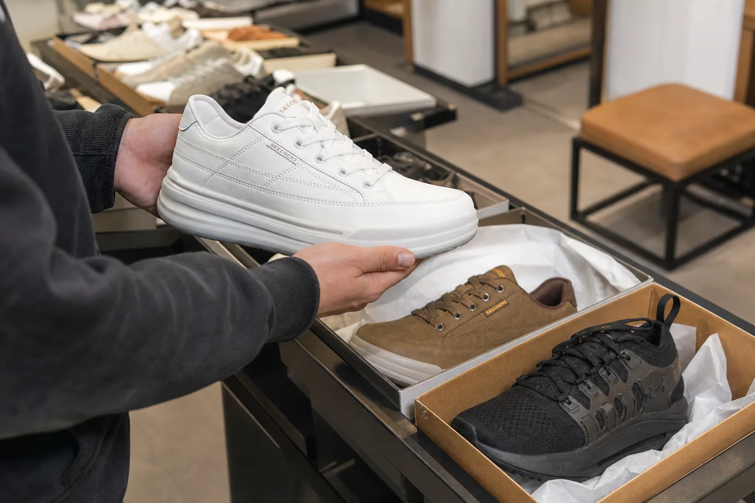 Outlet üzlet cipősorai között egy vásárló kézben tart egy fehér bőr sportcipőt, mellette nyitott cipősdobozok és különböző fazonú cipők sorakoznak egy alacsony polcon, a háttérben próbapad és tükör látszik.