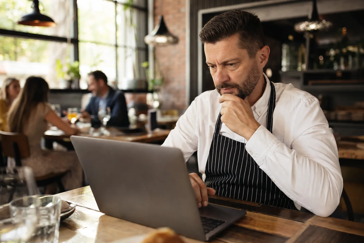 étteremvezető laptopon olvassa a Google értékeléseket, miközben háttérben vendégek étkeznek