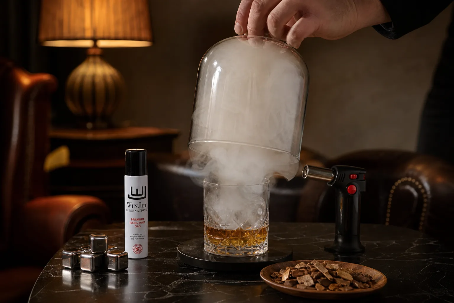 Esti otthoni nappaliban, dohányzóasztalon álló whiskeyspohár fölött üvegkupola alatt gomolygó sűrű fehér füst; a háttérben meleg fényű lámpa és karosszék látszik, az asztal mellett egy kéz éppen leemeli a kupolát, miközben a pohár mellett fa chips és kézi lángszóró hever.