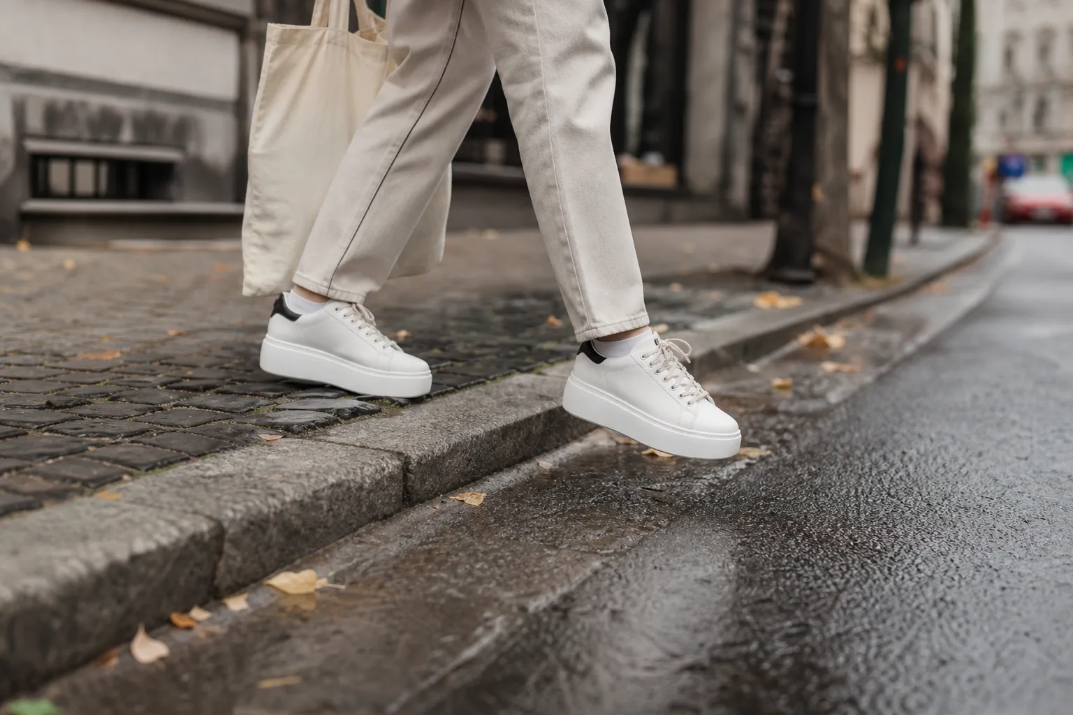 Városi járdán egy ember platform sneakerben lép le óvatosan egy alacsony padkáról, egyik kezében vászontáska, a közelben macskaköves rész és nedves aszfalt látszik, hogy a stabilitás és tapadás helyzetben legyen megfigyelhető.